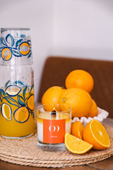 Candle with an orange label next to a glass with orange design and oranges on a woven mat.