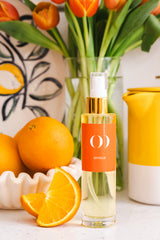 Glass bottle with orange label next to oranges and a vase of tulips on a white surface.