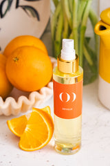 Bottle of Seville orange oil with oranges and a decorative shell on a white surface.