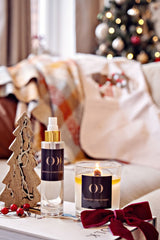 Decorative items including a candle, bottle, and wooden tree on a surface with a Christmas tree in the background.